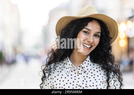 Magnifique jeune portrait de femme latino-américaine, femme marchant dans la ville du soir en chapeau avec des cheveux bouclés par temps chaud, souriant et regardant l'appareil photo de près. Banque D'Images