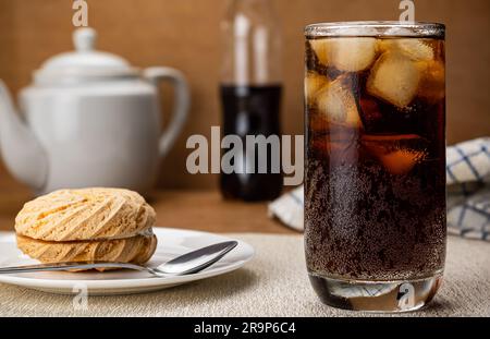 De l'eau froide au cola avec des glaçons en verre transparent et un délicieux petit gâteau rond fait maison rempli de crème aux myrtilles et d'une cuillère en métal de ceram blanc Banque D'Images