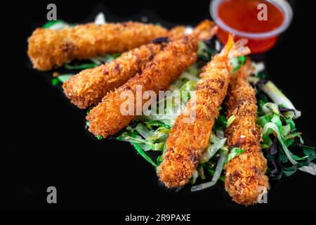 Crevettes enrobées de chapelure et frites sur feuilles de légumes et d'herbes avec sauce épicée sur fond noir, gros plan. Banque D'Images