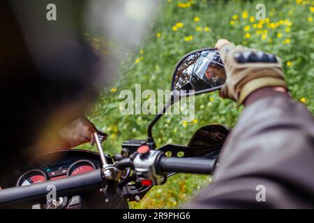 Motocycliste s'ajustant en regardant dans le miroir de moto à l'extérieur. Homme en cuir veste et casque dans la forêt d'été. Banque D'Images