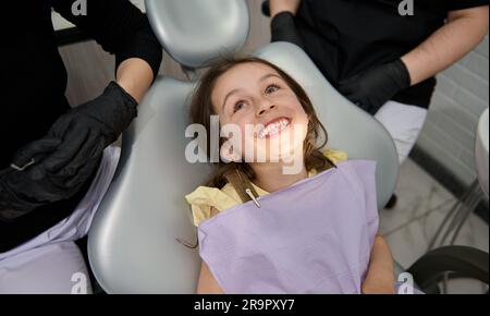 Petite fille mignonne assise dans une chaise de dentiste, souriant avec un beau sourire après le contrôle dentaire à la clinique dentaire Banque D'Images