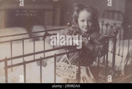 « At the Nest, Springfield » : carte postale photographique du début du 20th siècle. Il montre une jeune fille de l'Armée du Salut diriger la maison d'enfants 'The Nest' au 10 Springfield, Upper Clapton, Londres. Un panneau portant le nom « The Joy Leaf Cot » est accroché aux barres du lit de l'enfant. La carte postale a été envoyée en 1907. Banque D'Images