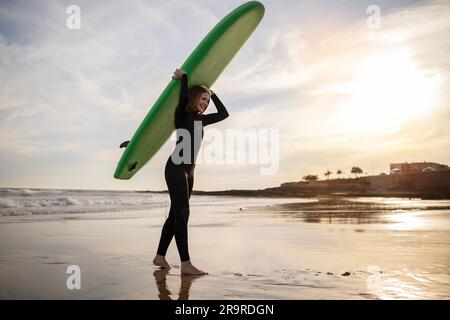 Portrait d'une jeune femme surfeuse marchant avec planche à voile à la plage au coucher du soleil Banque D'Images