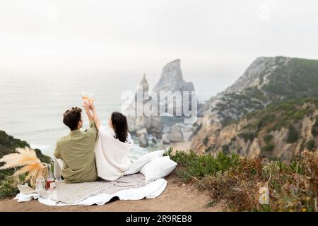 Jeune couple amoureux célébrant l'engagement ou la lune de miel sur la côte, en buvant du champagne, en regardant le bord de l'océan Banque D'Images