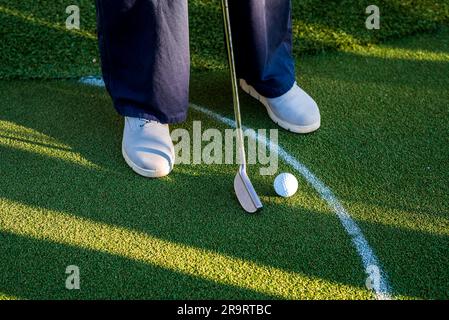 Mini-joueur de golf avec baskets grises mettant le ballon de golf dans le trou sur la voie verte. Banque D'Images