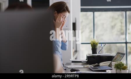 Employé de bureau traitant de la migraine et travaillant sur pc, frottant des temples et se sentant stressé. Femme d'affaires malheureuse ayant des maux de tête douloureux au travail, travaillant sous pression ou sous tension. Banque D'Images
