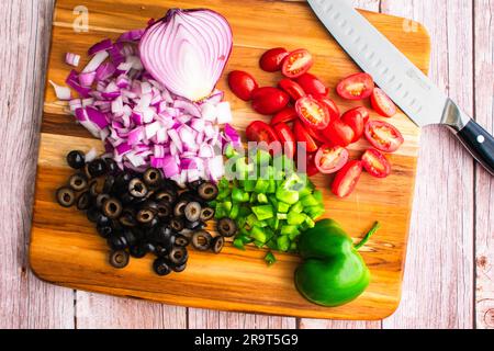 Poivron, tomates en raisin, oignon rouge et olives noires sur une planche à découper : légumes hachés et tranchés sur une planche à découper avec un couteau santoku Banque D'Images