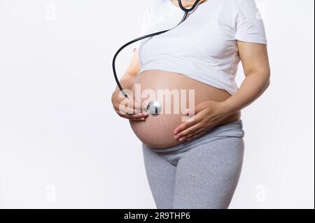 Une femme enceinte de près met un stéthoscope sur son ventre et écoute le rythme cardiaque de son bébé dans l'utérus. Développement du fœtus Banque D'Images