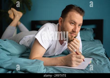 Homme millénaire posé sur le lit et écrivant un journal. Homme souriant et attentionné avec ordinateur portable et stylo dans ses mains. Banque D'Images