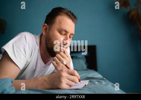 Homme millénaire posé sur le lit et écrivant un journal. Homme souriant et attentionné avec ordinateur portable et stylo dans ses mains. Banque D'Images