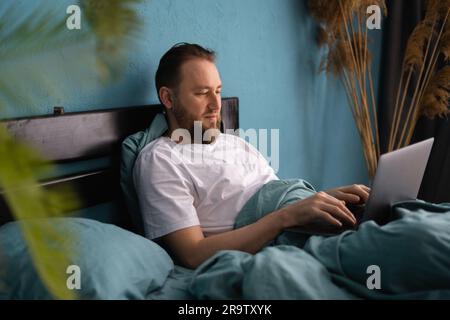 Homme barbu millénaire assis sur le lit de travail sur ordinateur portable dans la chambre à l'intérieur. Chat dans les réseaux sociaux, concept d'entreprise à la maison Banque D'Images