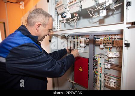 Un électricien en combinaison vérifie un blindage avec des fiches et des fils électriques. Banque D'Images