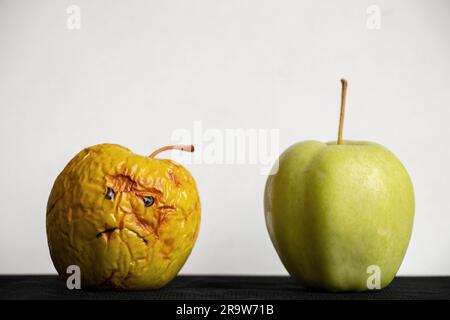 Une pomme pourrie et molle avec un triste sourire peint et une pomme verte mûre fraîche à côté sur un fond blanc sur une table noire, mauvaise et bonne appl Banque D'Images