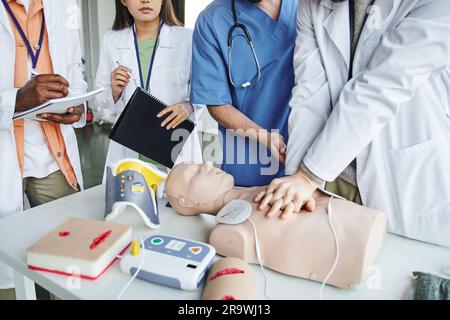 Vue rognée de l'homme afro-américain qui écrit dans un carnet à côté d'un professionnel de santé effectuant des compressions thoraciques sur un mannequin de RCP pendant un séminaire de premiers soins, Banque D'Images