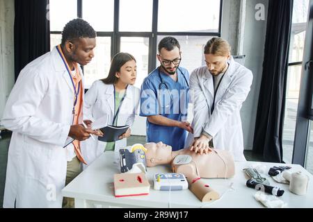 Étudiants multiethniques avec des cahiers regardant l'homme effectuant des compressions thoraciques sur le mannequin de RCP près du travailleur de santé et de l'équipement médical dans la formation ro Banque D'Images