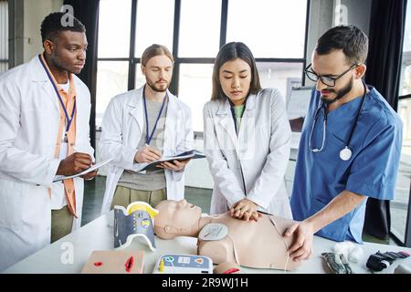 Femme asiatique pratiquant des compressions thoraciques sur un mannequin de RCP près d'un instructeur médical et d'étudiants interraciaux écrivant dans des carnets pendant la semina de premiers soins Banque D'Images