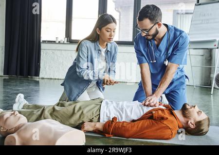 séminaire médical, professionnel de la santé en uniforme et lunettes montrant à une femme asiatique des techniques de réanimation cardiopulmonaire sur l'homme couché en formation Banque D'Images