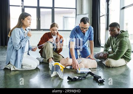 Professionnel paramédical montrant des techniques de réanimation cardiaque avec un mannequin de RCP et un défibrillateur aux participants multiculturels du séminaire de premiers soins, Banque D'Images