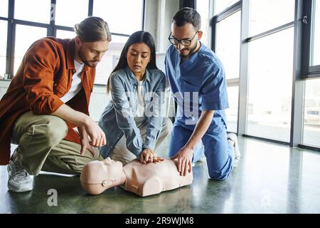 Femme asiatique pratiquant des compressions thoraciques sur un mannequin de RCP près d'un jeune homme et d'un instructeur médical en lunettes et uniforme pendant le séminaire de premiers soins, eme Banque D'Images