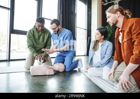 Instructeur médical expliquant les techniques de réanimation cardio-pulmonaire d'un homme afro-américain près d'un mannequin de RCP et d'une jeune équipe multiethnique, salvante s Banque D'Images