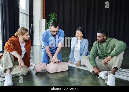 Professionnel paramédical effectuant des compressions thoraciques sur un mannequin de RCP tout en montrant des techniques de réanimation cardiopulmonaire à une jeune équipe multiculturelle duri Banque D'Images