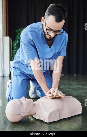 Jeune instructeur médical en lunettes et uniforme effectuant des compressions thoraciques sur le mannequin de RCP au sol dans la salle de formation, des compétences efficaces de sauvetage et Banque D'Images
