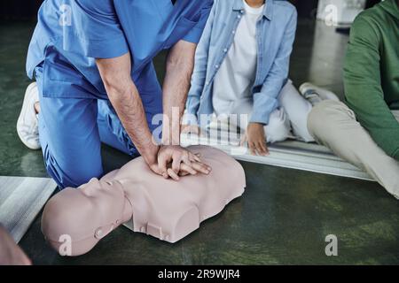 Réanimation cardiopulmonaire, vue partielle d'un instructeur médical effectuant des compressions thoraciques sur un mannequin de RCP, à proximité de jeunes participants au séminaire de premiers soins Banque D'Images