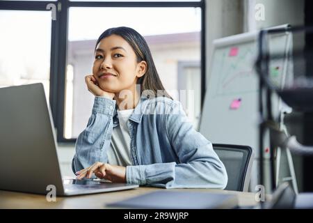 joyeuse femme d'affaires asiatique en chemise bleue denim, avec des cheveux de brunette assis près d'un ordinateur portable, tenant la main près du visage et regardant loin dans le bureau, jeune suce Banque D'Images