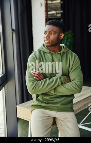 homme afro-américain pensif en vert élégant sweat à capuche assis sur un bureau avec les bras pliés et regardant loin dans le bureau moderne, jeune homme d'affaires, profession Banque D'Images