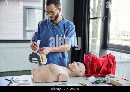 Professionnel ambulancier en uniforme bleu et lunettes tenant une étrésillon près du mannequin de RCP et du sac rouge lors de la préparation au séminaire de premiers soins, Life-savin Banque D'Images