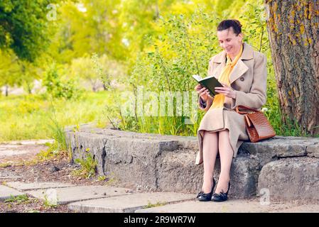 Femme élégante assise sur un mur de pierre au cours d'une journée de printemps ensoleillée, et la lecture d'un livre drôle Banque D'Images