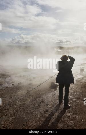 Un photographe photographiant dans la lumière du soleil et les embruns de Strokkur Geysir, à Geysir, dans le cercle d'Or, Islande. Banque D'Images