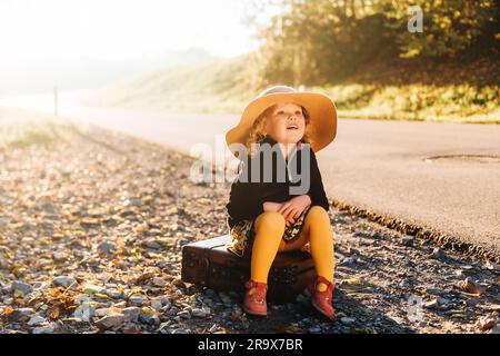 Portrait d'extérieur d'adorable petite fille adorable portant un grand chapeau, un manteau noir et des collants jaunes, assis sur une vieille valise vintage Banque D'Images