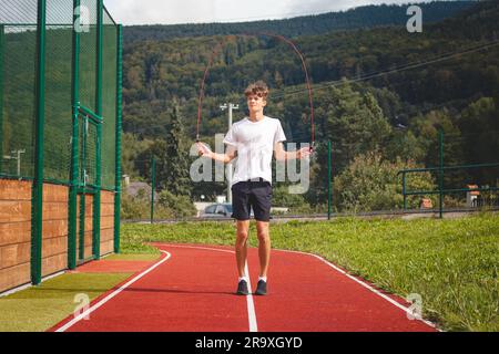 Un garçon aux cheveux bruns avec une silhouette athlétique portant un T-shirt blanc et un short noir est une corde de saut sur un ovale sportif. Entraînement pour améliorer le saut, co Banque D'Images