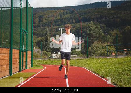 Un garçon aux cheveux bruns avec une silhouette athlétique portant un T-shirt blanc et un short noir est une corde de saut sur un ovale sportif. Entraînement pour améliorer le saut, co Banque D'Images