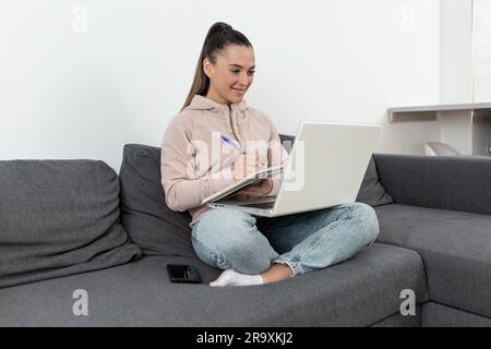 Jeune femme souriante dans des vêtements décontractés assis en position lotus sur le canapé à la maison, écrivant dans un ordinateur portable tout en tenant un ordinateur portable sur ses genoux Banque D'Images