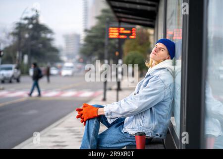 Un jeune homme fatigué touriste avec un café à emporter en attendant le bus. Homme blond sur l'arrêt de bus dans la ville Banque D'Images