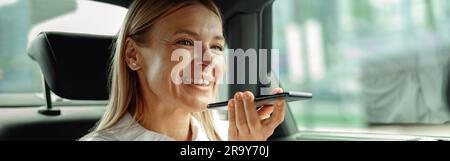 Femme d'affaires assise dans le siège arrière de la voiture enregistrant un message vocal sur le téléphone sur le chemin du travail Banque D'Images