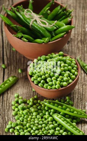 Gousses de pois verts et petits pois frais pelés dans des bols en céramique, grains de pois verts épars et gousses ouvertes sur fond de bois. Protéine végétale, pro Banque D'Images