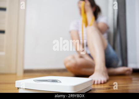 Femme en surpoids triste déprimé avec du ruban de mesure assis sur le sol près de l'échelle Banque D'Images