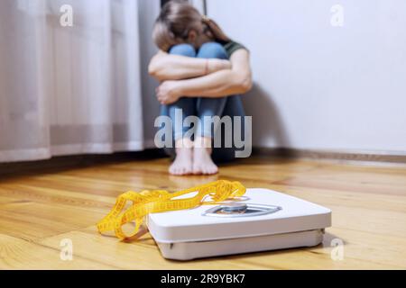 Femme triste et contrariée assise sur un sol en bois près de l'échelle blanche avec du ruban de mesure Banque D'Images