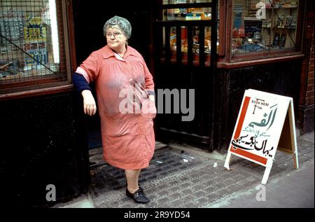 East End London 1970s, une femme anglaise plus âgée dans sa maison rouge debout sur une jambe et ses cheveux dans un filet à cheveux. Le panneau de la boutique écrit en partie en ourdou indique «Réservez ici pour Alif, école de l'automobile». Brick Lane, Whitechapel, Londres, Angleterre années 1978 70 Royaume-Uni HOMER SYKES Banque D'Images