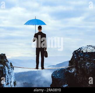 Risques et défis de l'entrepreneuriat. Homme d'affaires avec portfolio et parapluie debout sur corde au-dessus des abysses dans les montagnes, vue arrière Banque D'Images
