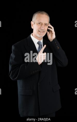 Un homme d'agréable portant un costume noir en souriant tout en parlant au téléphone mobile, sur fond noir Banque D'Images