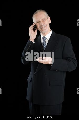 Un homme d'agréable portant un costume noir en souriant tout en parlant au téléphone mobile, sur fond noir Banque D'Images