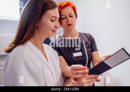 Jeune femme souriante regarde un catalogue de procédures et reçoit la consultation d'un dermatologue dans un salon de spa ou une clinique. Femme cosmétologue aux cheveux rouges Banque D'Images