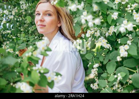 Gros plan d'un portrait d'une jeune belle femme blonde dans une chemise blanche avec des fleurs de jasmin. Une charmante fille sourit et pose à la caméra dans un ga Banque D'Images