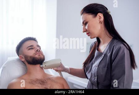 Jeune homme recevant une épilation photo lors d'une visite du centre de beauté dans un salon, esthéticienne professionnelle effectuant une procédure d'épilation laser sur le client Banque D'Images