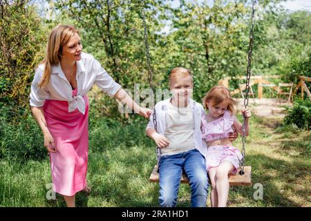 Une jeune maman attirante se déplace ses enfants sur une balançoire dans un pré dans le parc. Une femme blonde joue avec son fils et sa fille dans le jardin. Un heureux Banque D'Images