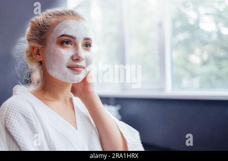 Jeune jolie fille avec un masque hydratant blanc appliqué sur son visage. Un blond aux yeux bleus agrémenté d'un peignoir blanc dispense des soins de spa dans un salon de beauté. Corps s Banque D'Images